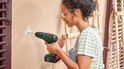 Vrouw met veiligheidsbril boort een gat in een gestucte muur met een groene Bosch accuboorschroevendraaier.