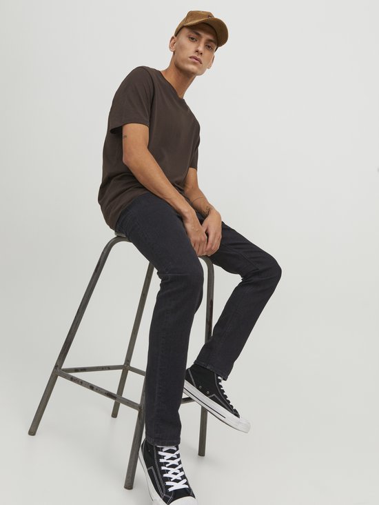 Man zittend op een kruk in bruine t-shirt, zwarte jeans, bruine pet en sneakers. Zwart denim.