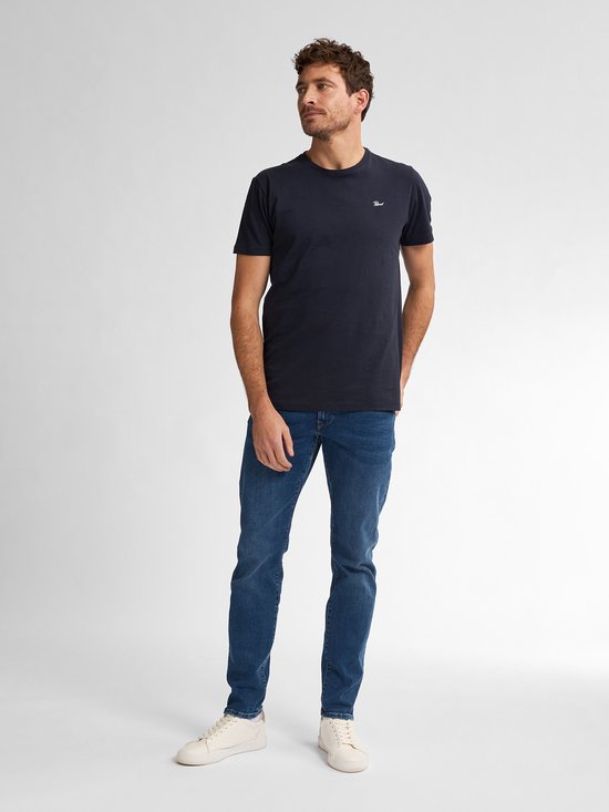 Man in donkerblauw T-shirt en blauwe spijkerbroek, staand.