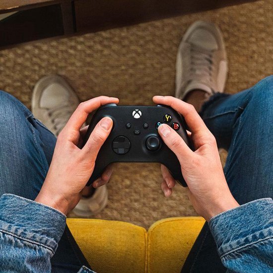 Persoon in spijkerbroek en spijkerjasje houdt een zwarte Xbox controller vast.