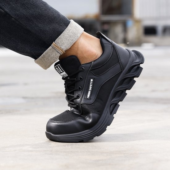 Zwarte werksneaker met robuuste zool, getoond aan een voet in een spijkerbroek.