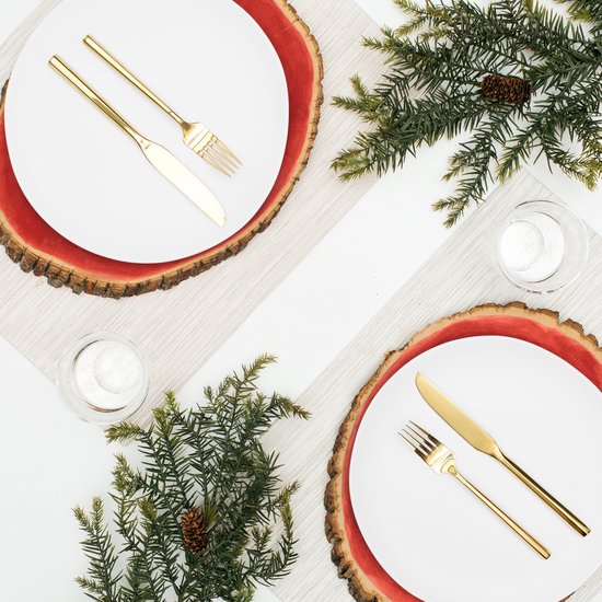Twee gedekte tafels met witte borden op houten onderzetters, gouden bestek, glazen en dennentakken.