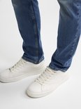 Gedetailleerde close-up van de onderbenen van een man, gedragen in een lichtblauwe spijkerbroek en witte sneakers.