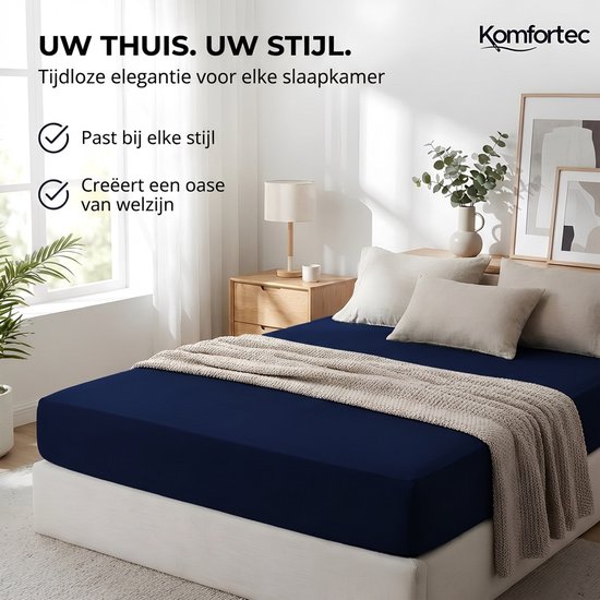 Een gedekt donkerblauw laken op een bed met beige kussens en plaid. Het laken is met een elastische band.