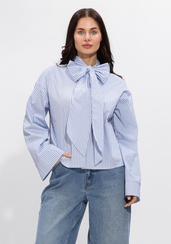 Vrouw met lange, bruine golvende haren, draagt een blauw-wit gestreepte blouse met strik en spijkerbroek.