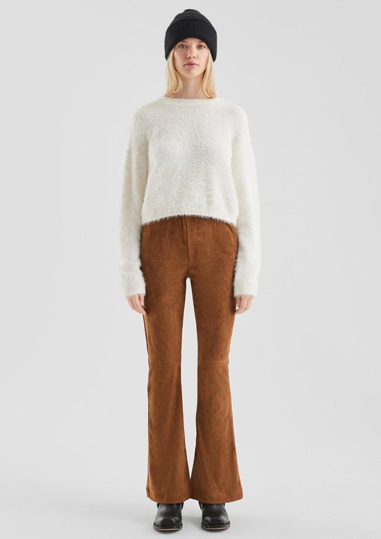 Model met witte, pluizige trui, bruine broek met split aan de onderkant en zwarte muts.