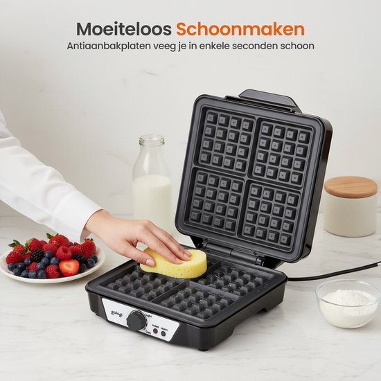 Een zwarte wafelmaker met open deksel, waar een hand een wafelplaat schoonmaakt met een gele spons. Naast de wafelmaker ligt een schaal met vers fruit.