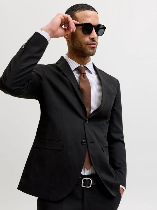 Man met zonnebril, gestreepte blouse, bruine das en zwarte blazer.