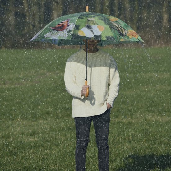 Un homme portant un pull jaune et un pantalon noir se tient sous un parapluie vert à motif de papillon sous la pluie.