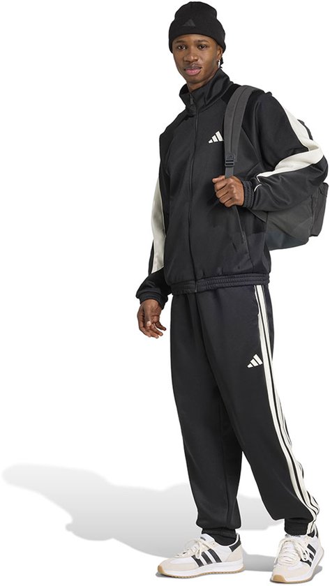 Man in zwart trainingspak met witte accenten, zwarte muts en rugzak, met zwarte trainingsbroek met witte strepen.