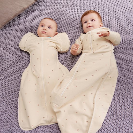 Twee baby's liggen op hun rug op een lila deken, beiden in crèmekleurige slaapzakken met geborduurde takjes.
