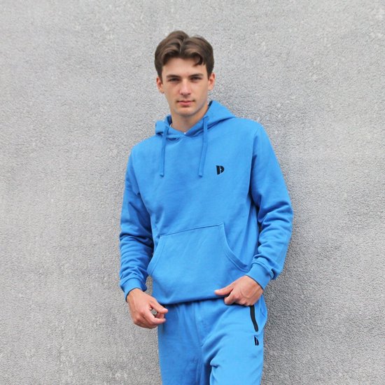 Jonge man in een blauwe hoodie en bijpassende joggingbroek, staand tegen een grijze muur.