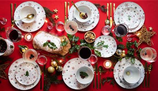 Kerstdiner setting met witte borden en kommen met kerstboom- en sneeuwvlokprint, gouden bestek, kaarsen en decoratie.