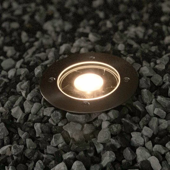 Verlichte ronde grondspot in een bed van donkere stenen. De lamp geeft een zachte, warme gloed.