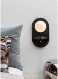 Zwarte wandlamp met ronde melkglazen kap en schapje, naast een bed met blauw en gestreept kussen.