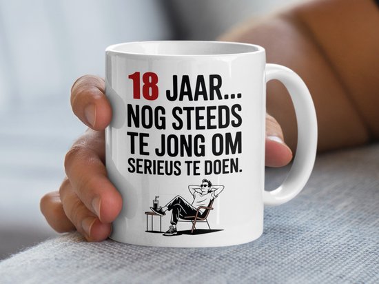 Witte mok met tekst "18 JAAR... NOG STEEDS TE JONG OM SERIEUS TE DOEN." en illustratie van een relaxte man in een stoel.