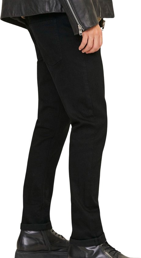 Zij-aanzicht van een model in zwarte jeans, bruine coltrui en leren jack met zwarte laarzen.