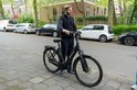 Man op een zwarte Sparta e-bike, staand op een stoep, met auto's op de achtergrond.