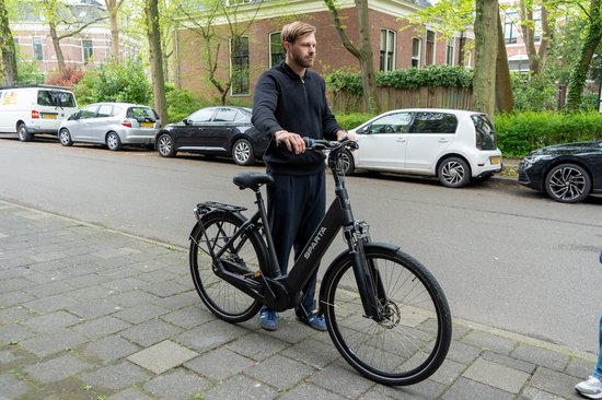Man op een zwarte Sparta e-bike, staand op een stoep, met auto's op de achtergrond.