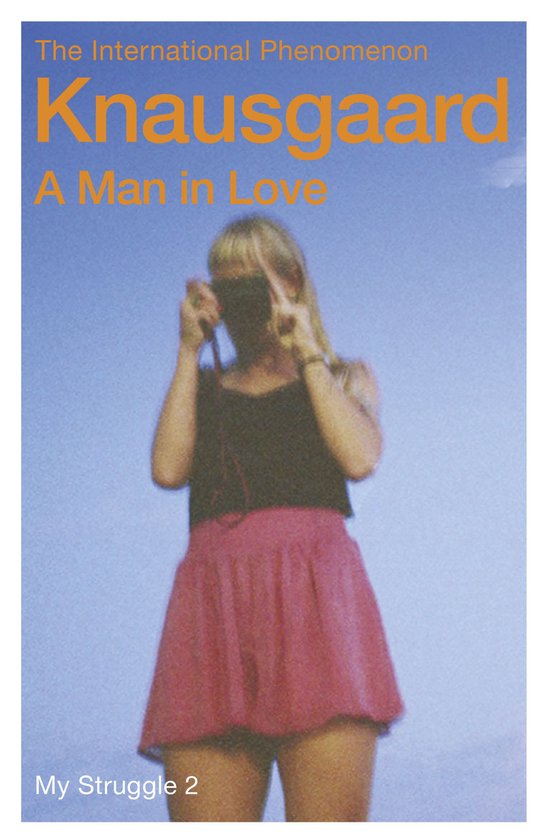 Een vrouw met blond haar draagt een roze rok en een zwart topje, terwijl ze een camera vasthoudt tegen een blauwe lucht. Knausgård: A Man in Love.