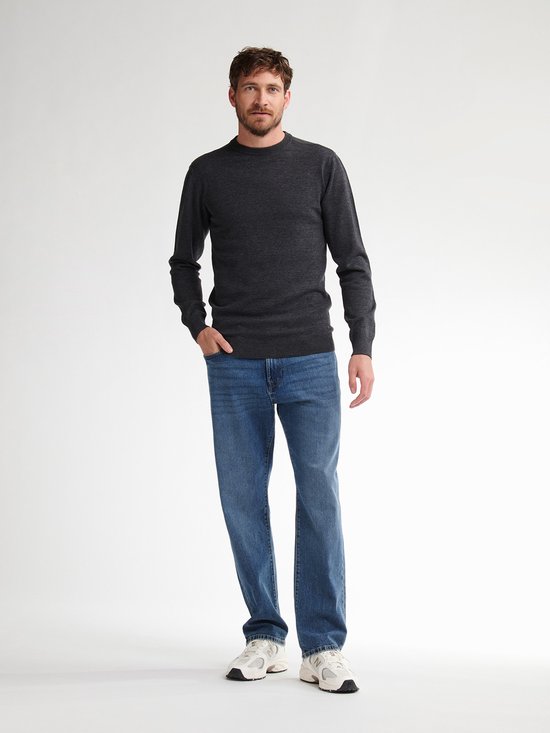 Man staat frontaal in een donkergrijze gebreide trui en blauwe jeans.