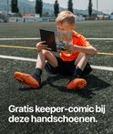 Jongen zit op veld en leest comic "BE THE TITAN". Draagt oranje shirt en voetbalschoenen.