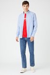 Man, staand, draagt een blauwe spijkerbroek met een rode Wrangler t-shirt en blauw overhemd.