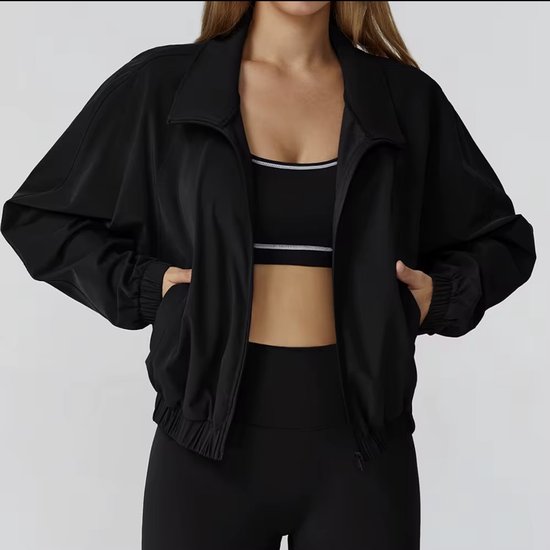 Model draagt een zwarte sportbeha en legging, met daarboven een loszittende zwarte jas. De jas heeft een rits aan de voorkant.