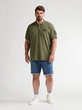 Man met baard van voren gezien, draagt olijfgroene polo met opdruk en blauwe denim shorts met opgerolde zoom. Draagt witte sokken met zwarte strepen en witte sneakers.