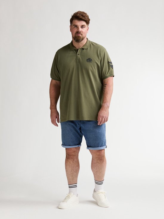 Man met baard van voren gezien, draagt olijfgroene polo met opdruk en blauwe denim shorts met opgerolde zoom. Draagt witte sokken met zwarte strepen en witte sneakers.
