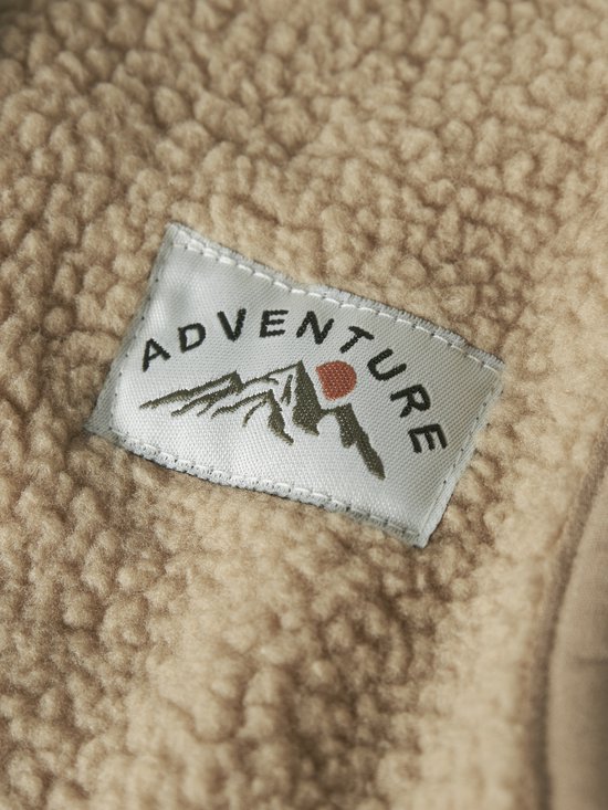 Detailopname van beige teddy-stof met een geweven label met de tekst 'ADVENTURE' en een bergillustratie.