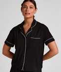 Model draagt een zwart pyjamashirt met korte mouwen en witte piping. Het shirt heeft een open kraag en een borstzakje.