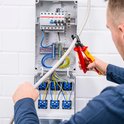 Elektricien met rode en gele kniptang knipt door een grijze kabel, terwijl deze is aangesloten op een schakelkast.