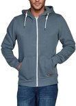 Man in blauwgrijze hoodie met rits, spijkerbroek en handen in de kangoeroezakken.