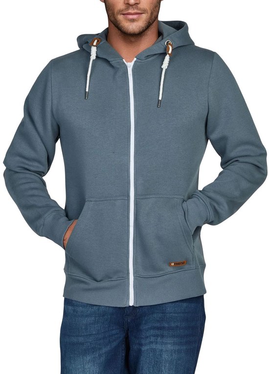 Man in blauwgrijze hoodie met rits, spijkerbroek en handen in de kangoeroezakken.