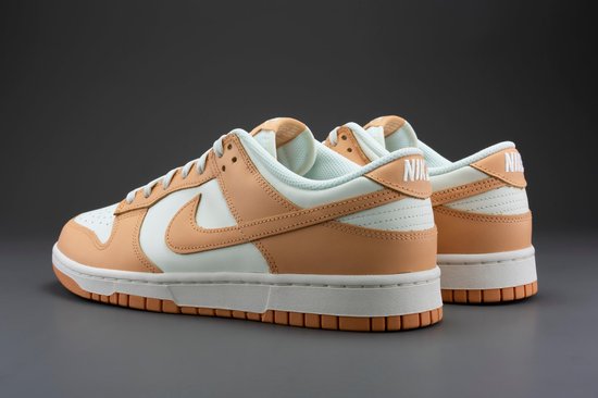 Een paar Nike Dunk Low damesschoenen, wit met oranje overlays, op een grijze achtergrond.