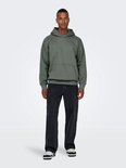 Man van voren, met een groene hoodie en wijde, zwarte spijkerbroek met stonewashed look en sneakers.