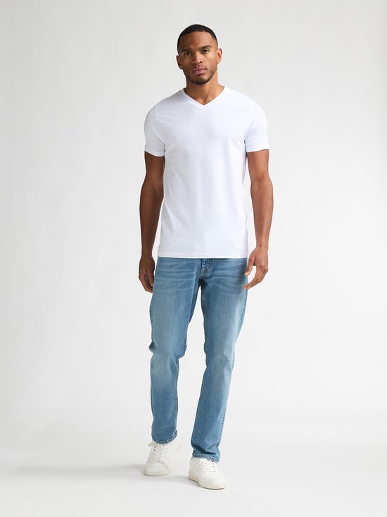 Man met korte zwarte haren, met baardje, draagt een wit V-hals T-shirt en een spijkerbroek.
