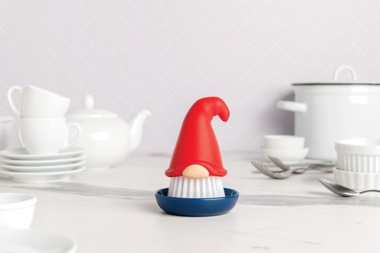 Een afwasborstel in de vorm van een gnome met rode puntmuts, beige neus en witte borstelharen, staand in een donkerblauwe houder, op een aanrecht met keukengerei.