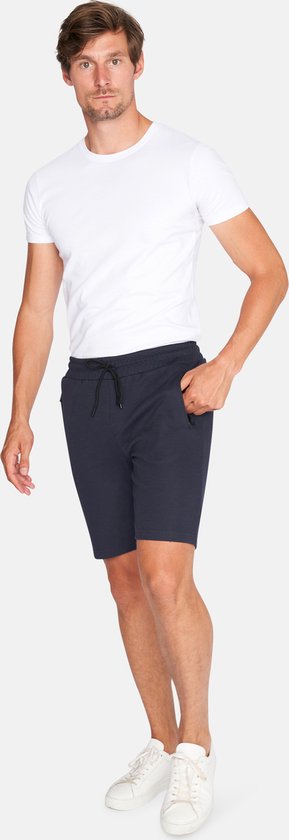 Man met witte t-shirt en donkerblauwe korte broek met trekkoord en ritssluitingen aan de zijkant, staand.