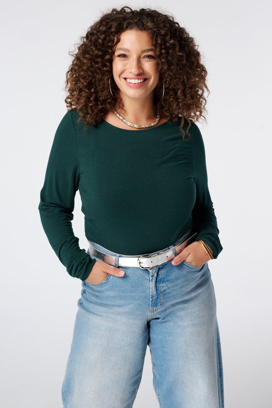 Model draagt een donkergroene top met lange mouwen, wijde spijkerbroek en een zilveren riem.