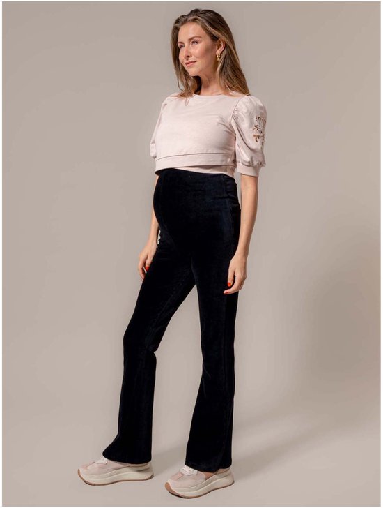Zwangere vrouw draagt een zwarte corduroy zwangerschapsbroek met uitlopende pijpen en een beige top.