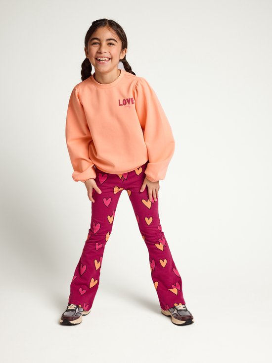 Meisje draagt een perzikkleurige sweater met 'LOVE' opdruk en een bordeauxrode flared broek met hartjes.