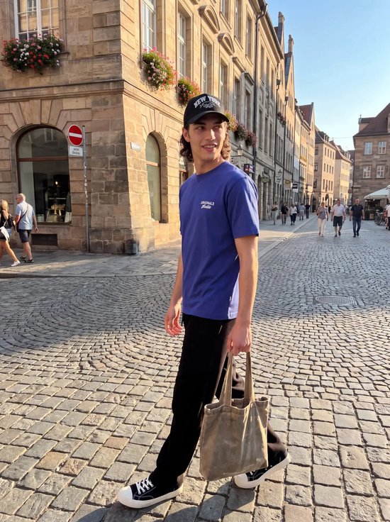 Model loopt over een geplaveide straat in een blauw T-shirt, zwarte pet en zwarte joggingbroek met een draagtas.