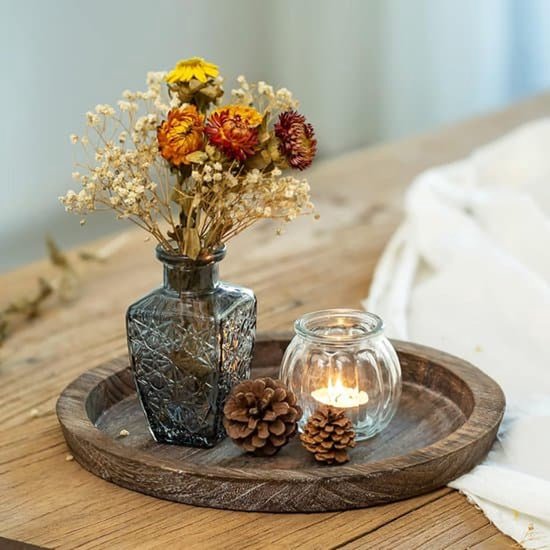 Rond houten dienblad met een glazen vaas met droogbloemen, een glazen theelichthouder en kleine dennenappels.