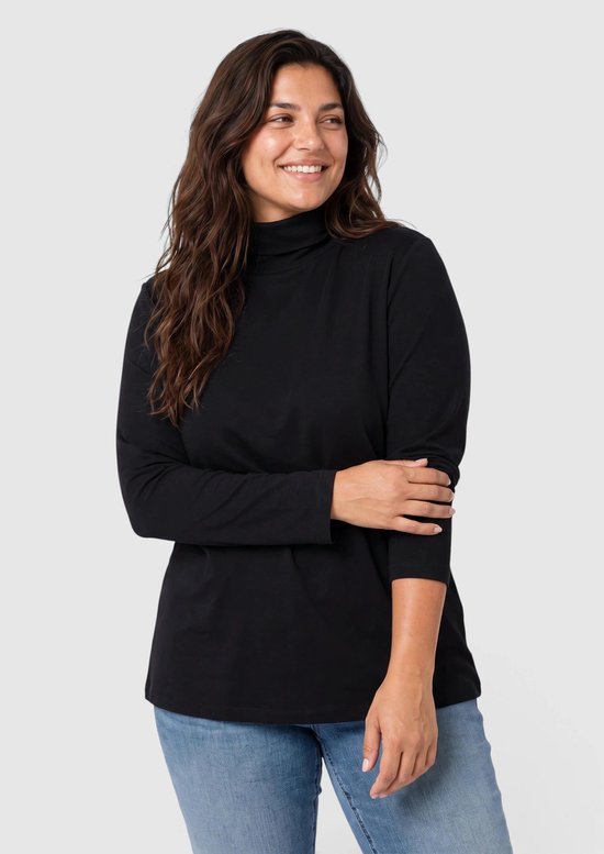 Vrouw met lang donkerbruin haar draagt een zwarte coltrui met lange mouwen en een jeans.