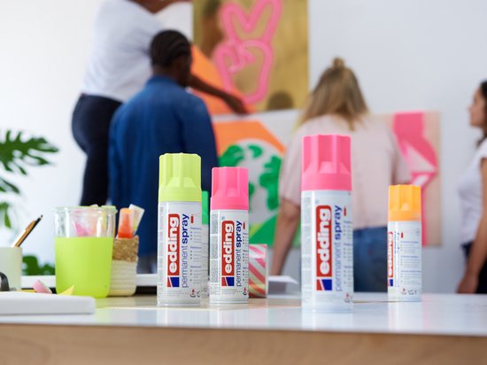 Bouteilles de spray néon rose, vert vif et rose d'edding sur une table, avec des œuvres d'art en arrière-plan.