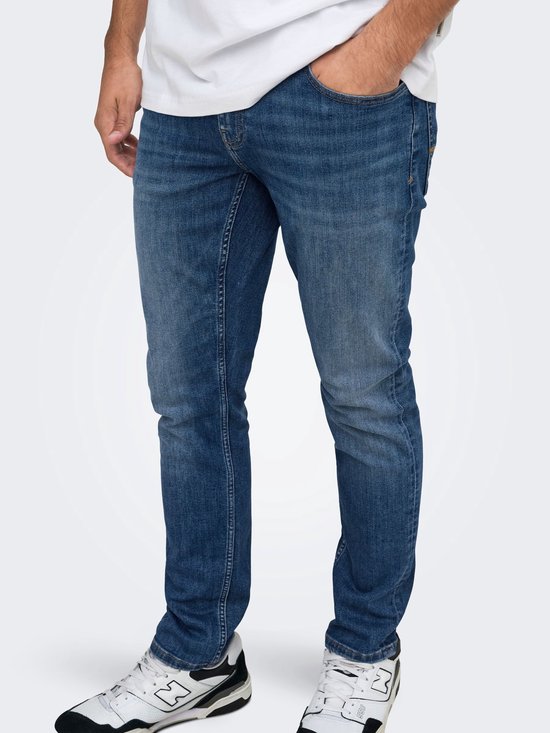 Close-up van de pasvorm van een donkerblauwe slim-fit jeans op een model, gecombineerd met zwart-witte sneakers.