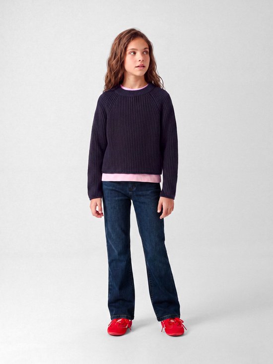 Meisje draagt een donkerblauwe bootcut jeans met een roze shirt, een donkerpaarse trui en rode sneakers.
