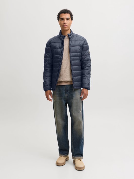 Model draagt een donkerblauw, gewatteerd pufferjack met opstaande kraag, gecombineerd met een beige trui en jeans.